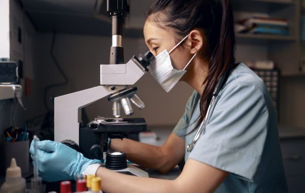Asian woman with medical mask looks into microscope at table in hospital