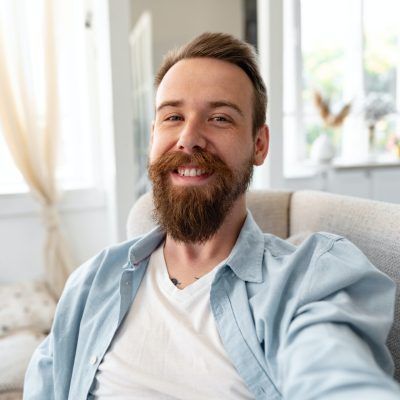 Bearded happy young man smiling and taking selfie photo while sitting on sofa at home.