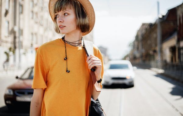 Young girl with headphones in a yellow T-shirt and a straw hat walks with a backpack along a city